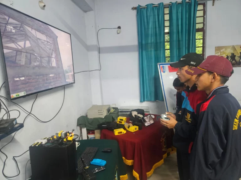 Trainers demonstrating FPV drone equipment beside a screen showing live drone footage at the Gangtok NCC workshop